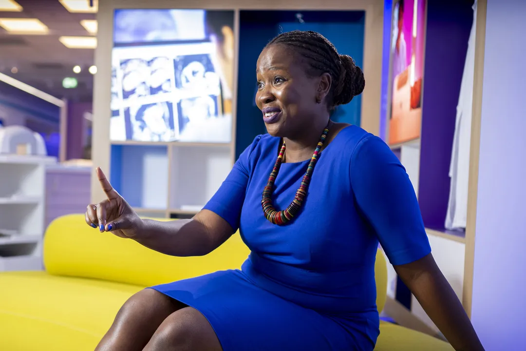 Black woman smiling wearing smart blue dress and beaded necklace sitting on a yellow sofa in an office