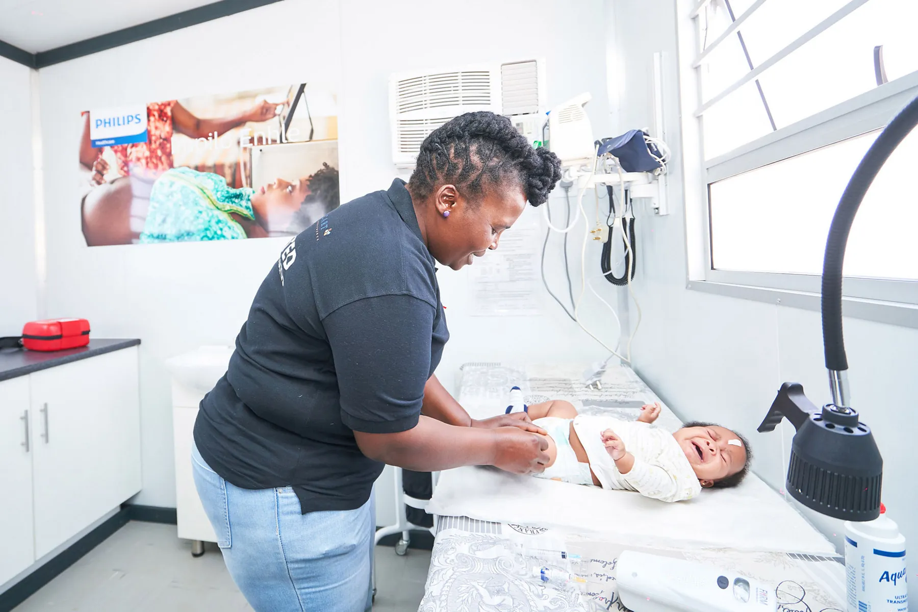 Black female clinician in black polo shirt examining a crying baby on a hospital bed next to a window