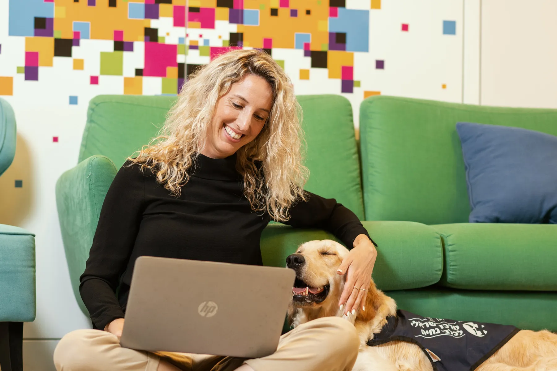 Female office worker sat against a green sofa with a laptop and a labrador dog