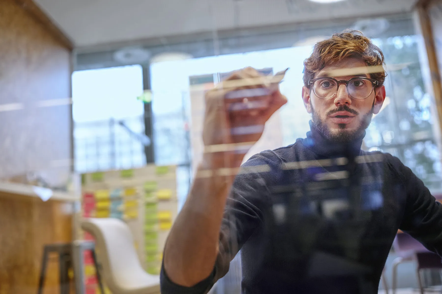 Front view of a young bearded technology professional wearing glasses writing ideas on a glass screen