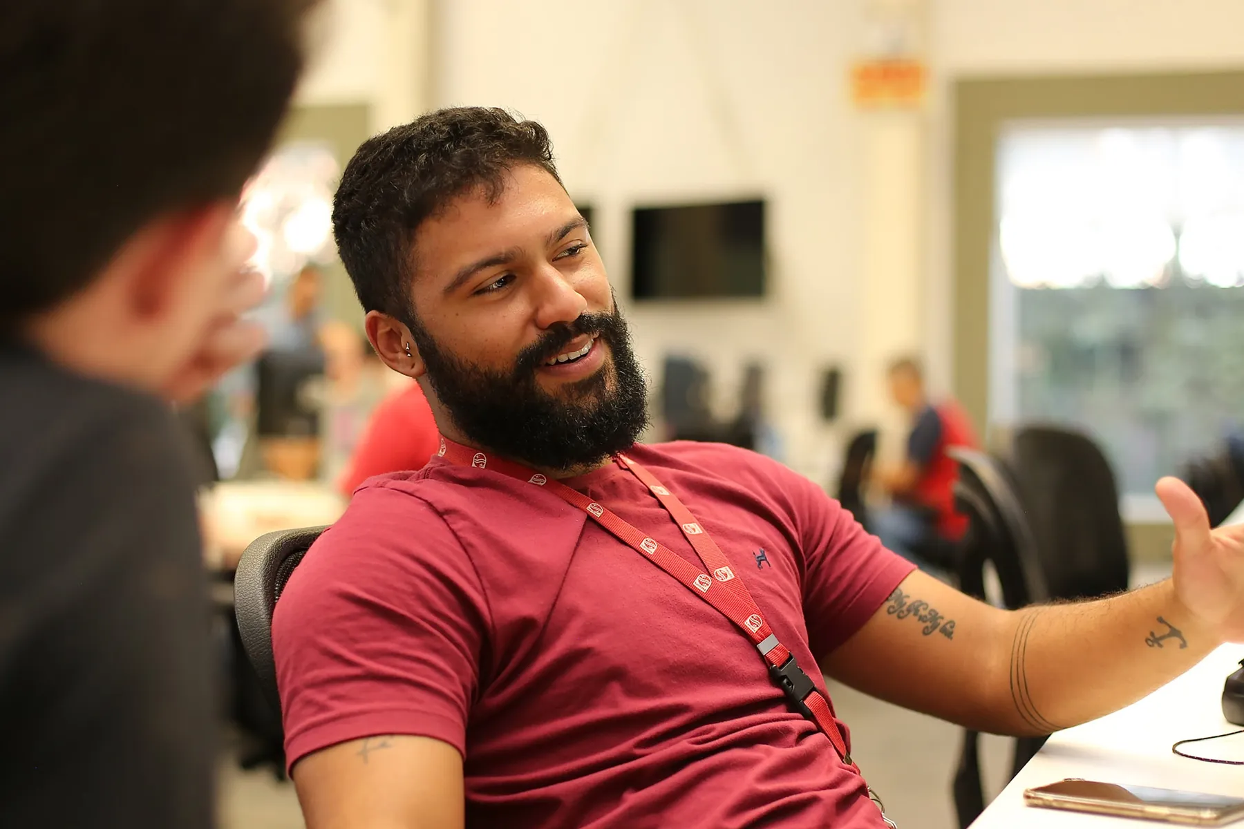 Young male office worker wearing red Philips lanyard around his neck and a red t-shirt sat in an office chair speaking to a colleague