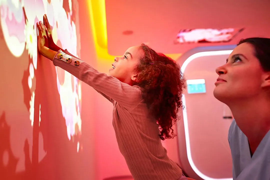 Female nurse behind young girl reaching up and touching a projected image on a wall in a medical room with soft pink lighting