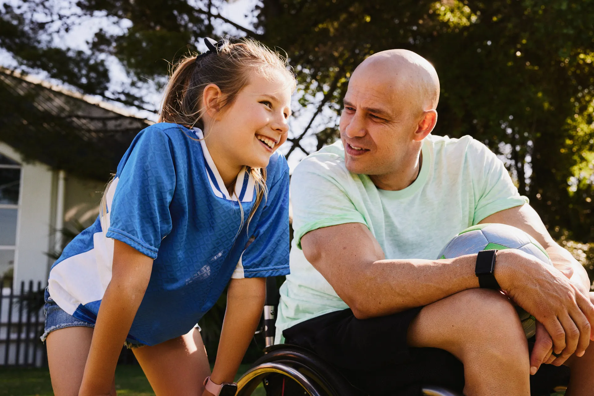 Young girl smiling next to athletic bald man in wheelchair holding a soccer ball